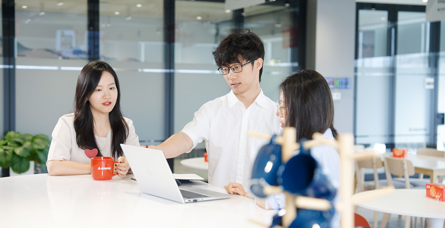 Three people standing around a table looking at a laptop in the cafeteria of an office