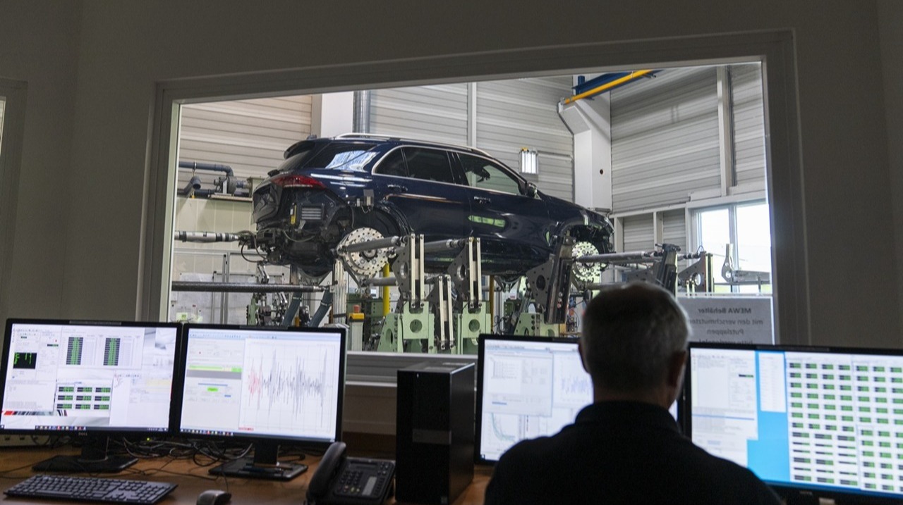 Engineer monitoring data on multiple computer screens while a car undergoes testing in a lab through a large observation window