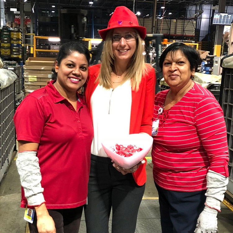Adriana Huntington wearing a red cowboy hat and coworkers wearing red shirts and holding candy bowl filled with chocolates. In a manufacturing facility.