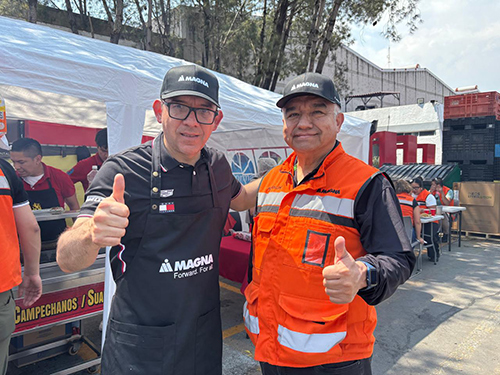 Miguel Arellano and other person in branded attire give thumbs up at an outdoor event, smiling warmly. One in a black apron, the other in an orange vest. Casual, friendly atmosphere.