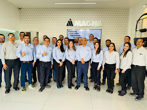A group of 17 Magna Employees stand smiling and posing in front of a "MAGNA" sign. They wear matching uniforms, creating a professional and positive atmosphere.